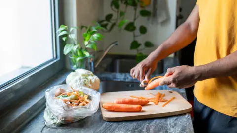 A person peels carrots over a wooden chopping board. They have a bag next to them with the peelings in and with other food waste. In the background there is a sink and plants.
