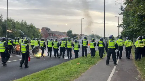 A line of police in riot gear stand across a road as fireworks are launched at them.