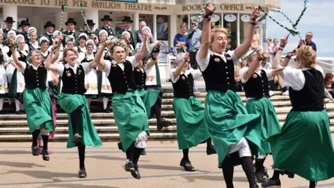 A troupe of seven female clog dancers in long green skirts, black waistcoats and white short-sleeved collared shirts. They are jumping with their arms in the air in front of wide steps leading up to the entrance to a pleasure pier. More clog dancers are standing on the steps, in different costumes.