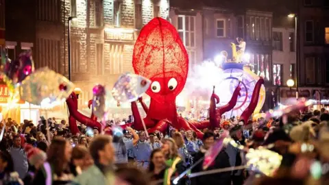 A street party in Morecambe. Hundreds of people are seen along the street whilst a large shrimp puppet is held up.