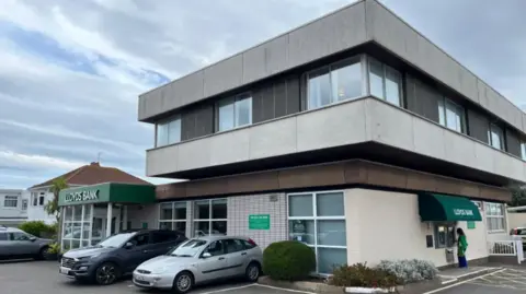 Lloyds Bank branch in Les Quennevais, Jersey, which has several cars parked in its car park and a woman using one of the two ATM machines.