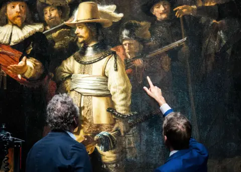 Getty Images Two visitors in front of Rembrandt's The Night Watch. The visitor on the right is pointing at lieutenant Willem van Ruytenburch in the painting, who is dressed in a gold outfit with a white sash.