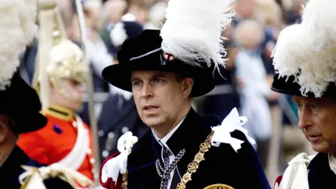 Michael Dunlea/PA Media Prince Andrew in the regalia of the Order of the Garter. He is wearing a black jacket and hat with gold chains, white ribbons and a white feather from his cap. The King, wearing the same regalia, can be seen to his left