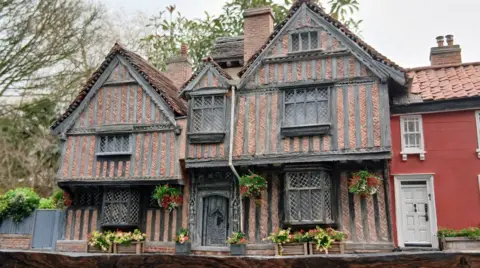 Simon Evans A model of a medieval building in a brown colour. It has a wooden door and there are several windows placed across three-storeys. There are model plant boxes and hangers on the front of the home.