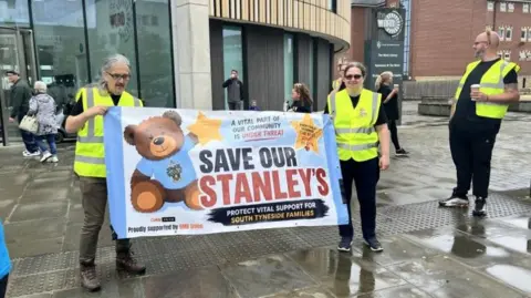 Two people wearing hi viz vests holding a banner between them. The man on the left has tied-back grey hair and is wearing spectacles and brown trousers and boots. To the right the woman has dark tied-back hair and is wearing sunglasses and dark trousers. The banner features an image of a teddy bear in a blue t shirt and holding a star, and its slogan states: Save our Stanley's. To the left a man also in a hi viz vest, and wearing dark clothing is holding a cup and looking towards the building behind them which has a rounded glass entrance.