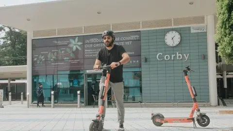 Voi/North Northamptonshire Council A man getting on an e-scooter outside Corby Railway Station - a mostly glass fronted building, with a clock to the right. Another red e-scooter is to the right. The man is wearing a black T-shirt and grey trousers, a helmet and sunglasses. He also has a beard.