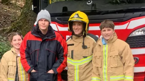 Four people in front of a red fire engine. Two are children in beige firefighter's coats and there is also a woman in a beige firefighters coat and yellow helmet alongside a man in a blue and red coat and get beanie hat