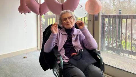 Dorothy Howard smiles for a photo in her wheelchair. She s wearing a light purple cardigan over a dark purple roll neck jumper. She is wearing glasses and has her hands raised in celebration. Pink balloons spelling '100' can be seen in the background.