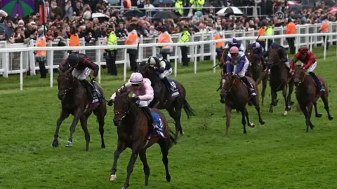 Eight competitors competing at a horse racing event. There is a line of security staff, in orange and yellow hi-vis jackets, standing in front of a crowd. 