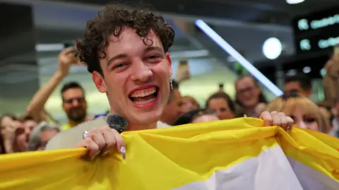 Last year's winner Nemo holding a non-binary flag and smiling to the camera at Zurich airport. 