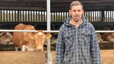 Josh Dorey is smiling at the camera as he stands outside a cattle shed. Behind and to the side of him is a Guernsey cow who is poking her head through a bar fence and is looking directly at the camera. There are several other cows lying down inside the shed. Mr Dorey has short brown hair and a beard and is wearing two layers under a thick, checked and hooded shirt.  