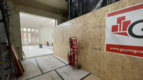 Shaun Whitmore/BBC The photo shows a part of the museum and an adjoining room. Construction materials can be seen resting along the walls while a protective surface covers the flood. A bucket can be seen in the background along with several windows.