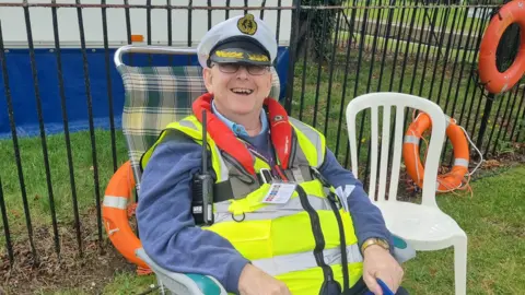 Martyn Pearson, dressed in a naval outfit, peaked cap and high-vis jacket, is sitting in a chair and smiling. He is in front of a black, metal, railed fence and there is a white plastic chair next to him. He is also wearing a blue jumper and has a walkie talkie on a strap around his chest.