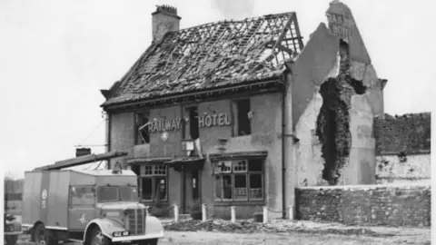 An exterior shot of the former Railway Hotel in Catterick, North Yorkshire, after an ammunition explosion in 1944. The roof is destroyed and there is a large hole on the right hand side wall. Parked outside is a fire engine. 