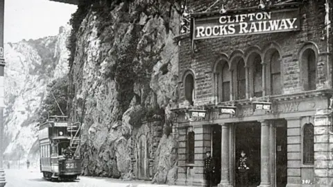 Clifton Rocks Railway An archived black and white image of the Clifton Rocks Railway in operation. There is a large sign hanging above the tunnels and two members of staff are standing in the doorway. A tram car is driving past on what is now the Portway road.