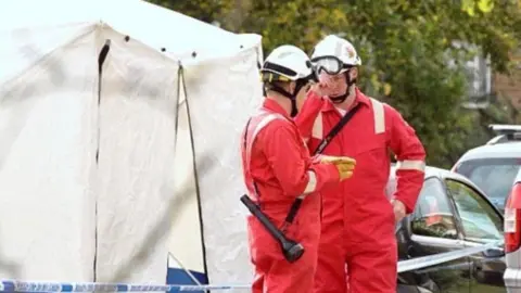 David Hadjicostas Two fire officers dressed in red overalls and white helmets stand talking to each other. In the background is a white forensic tent and a number of cars can bee seen parked behind it.