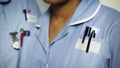 A stock image of two nurse's torsos, both wearing blue uniforms, with pens in breast pockets.