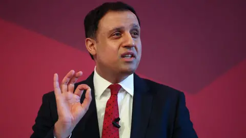 Getty Images A man with short black hair is wearing a black suit jacket, white shirt and red tie. He is standing in front of a red backdrop
