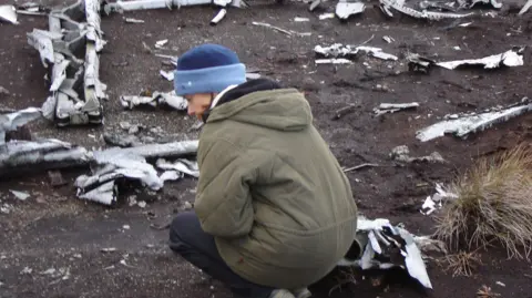 Carol Kyle-Sage A woman kneels down at the Cheviots crash site. She is wearing a blue hat and a khaki green thick coat. Debris from the B17 Bomber can be seen scattered on the hills. 