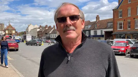 Gary Farrow. He is pictured in Marlborough town centre. He has short grey hair and a grey moustache. He is wearing dark square shaped sunglasses. 