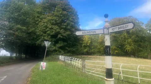 Photograph of a road sign on the edge of the village of Adlington in Cheshire. It is a rural road, with trees and farmland in the background.