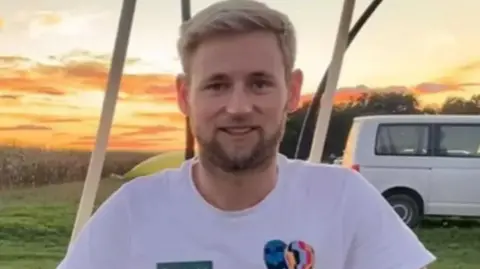 A man smiling towards the camera wearing a white tshirt with a mini hot air balloon graphic on the right hand side. He is sitting in a field at sunset, with a van behind him