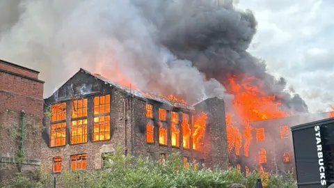 Kiyan Beasant An old brick building on fire. Huge orange flames are inside the building and burning out the windows all around it. Thick black smoke is coming from the fire.