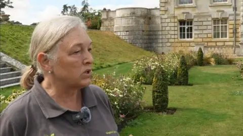 BBC/Sophie Cridland Clare Gascoigne stand in the formal gardens at Kingston Lacy, standing beside flower borders and lawns in front of the historic stone mansion. Parts of the grass appear dry and yellowed, while flowering plants line the garden beds. The house's ornate façade, windows and terrace can be seen in the background.