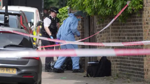 PA Media A forensic investigator in a blue suit and face mask examines a scene behind police tape, with a uniformed officer and police vehicles nearby.
