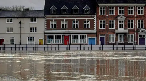 Simon Beckett The River Severn, in flood, passes through Bewdley, with grey flood defences in operation and buildings behinds. Two are red-brick with white window frames, and the third is an olive colour with a yellow door