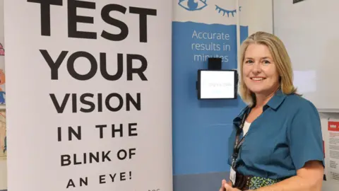 Dr Louise Allen is standing in front of a large sign that says "test your vision in the blink of an eye". There is a tablet on the wall and another sign that says "accurate results in minutes". Dr Allen has shoulder length fair hair and is smiling at the camera. She is wearing a blue blouse and has a name tag around her neck