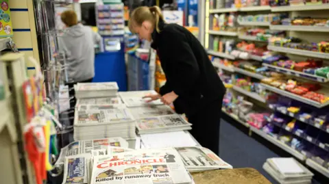 A woman sorts through a pile of newspapers stacked on a table in her newsagents. Behind her are display shelves containing confectionery.