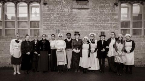 The present day teachers of Lacock Primary School lined up against the wall in Victorian outfits