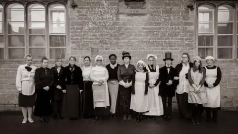 The present day teachers of Lacock Primary School lined up against the wall in Victorian outfits