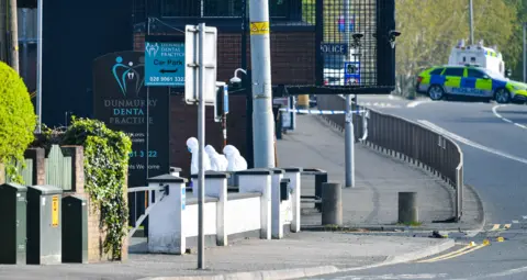 Pacemaker Three people in white forensic suits examine the scene outside a police station. A police car with a blue and yellow checked pattern can be seen in the background and there is blue and white police tape tied to a lamppost