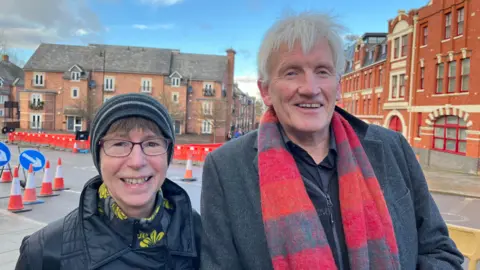 Jo and Kevin stood side by side, both wearing winter coats, hats and scarves. Behind them you can see the gyratory roads, with lots of bright orange traffic cones, orange barriers, and blue signs to help drivers. 