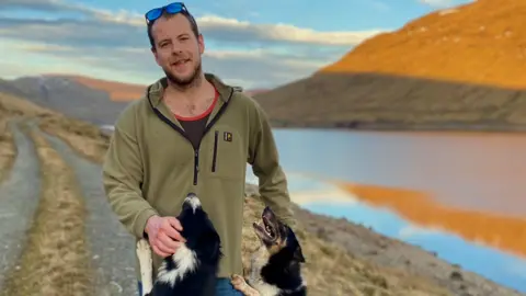 Netflix Man wearing sunglasses on his head smiling at the camera with two black and white dogs jumping up at him 