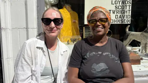 BBC/Chloe Hughes Two women are smiling - one has blonde hair, a white shirt and sunglasses and the other has an orange headscarf, a black t shirt and a big gold hoop earrings. 