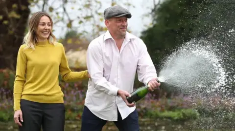 A man in a white shirt and cloth cap opening a bottle of champagne outdoors with spray everywhere next to a woman in a yellow jumper.