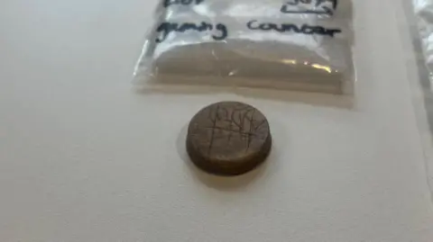 Uncovering Roman Carlisle A round gaming counter. It is dark brown in colour and has some carvings on it. Above it is visible part of a plastic bag used to preserve it.
