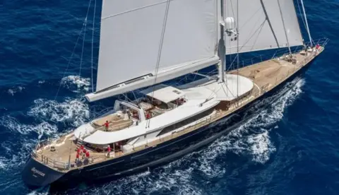 Perini Navi Press Office A large luxury yacht can be seen sailing through water with large white sails unfurled and crew members in red uniform on the deck