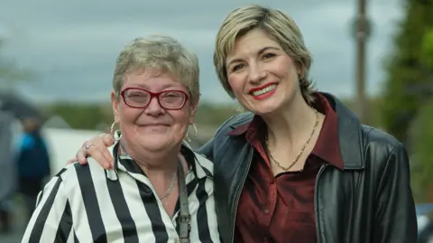 James Stack/Netflix The real Susan McIntyre is standing with Jodie Whittaker. Both are smiling and Jodie has an arm around Susan.