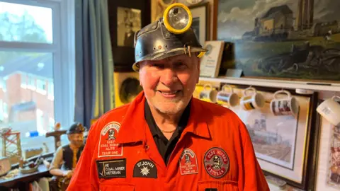 A man wearing orange mining overalls and a helmet with a torch on it stands in a room full of mining memorabilia with pictures on the wall.