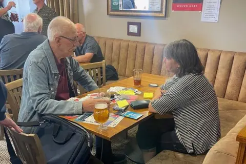 Karen Goddard Karen Goddard interviewing a dock worker. They are sitting opposite each other at a brown table. On the table are drinks, notes and a tape recorder
