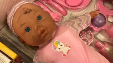 BBC A baby doll in a pink outfit has soot marks on its cheek, nose and forehead. Its surrounded by other pink coloured toys. 