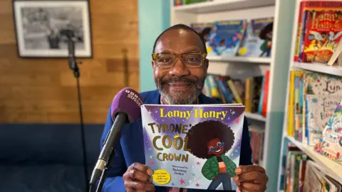 Sir Lenny Henry smiling at the camera sitting inside Afrori Books in Brighton holding his latest book Tyrone's Cool Crown