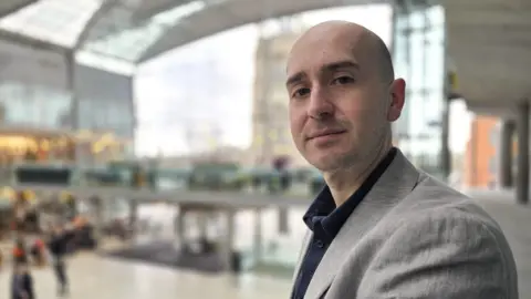 Ben Goldsborough is looking at the camera. He's wearing a grey suit jacket over a dark shirt, and is standing in the Forum in Norwich.