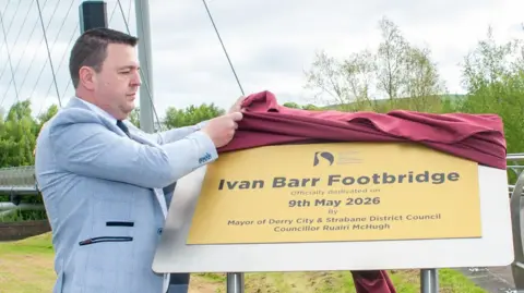 Mayor of Derry City and Strabane District Ruairí McHugh, who has short brown hair and is wearing a blue suit, is seen pulling a red cloth over a golden plaque with the wording: 'Ivan Barr Footbridge'. 