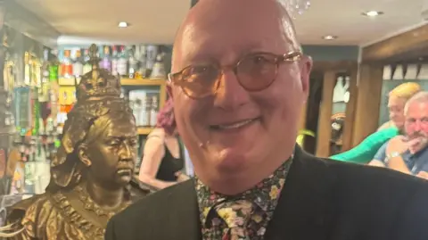 James Burrell-Cook James Burrell is wearing a floral shirt and a florla tie with a black blazer. Behind him in the pub is the gold bust of Queen Vistoria.