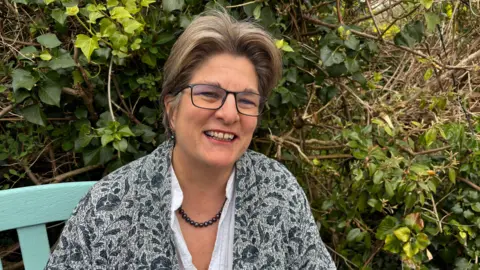 A woman in glasses and a floral cardigan sitting on a bench